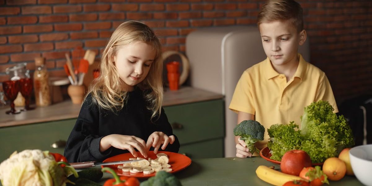 Positive little girl and boy sitting at desk with various fresh vegetables and fruits while preparing meal in loft kitchen with brick wall