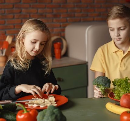 Positive little girl and boy sitting at desk with various fresh vegetables and fruits while preparing meal in loft kitchen with brick wall