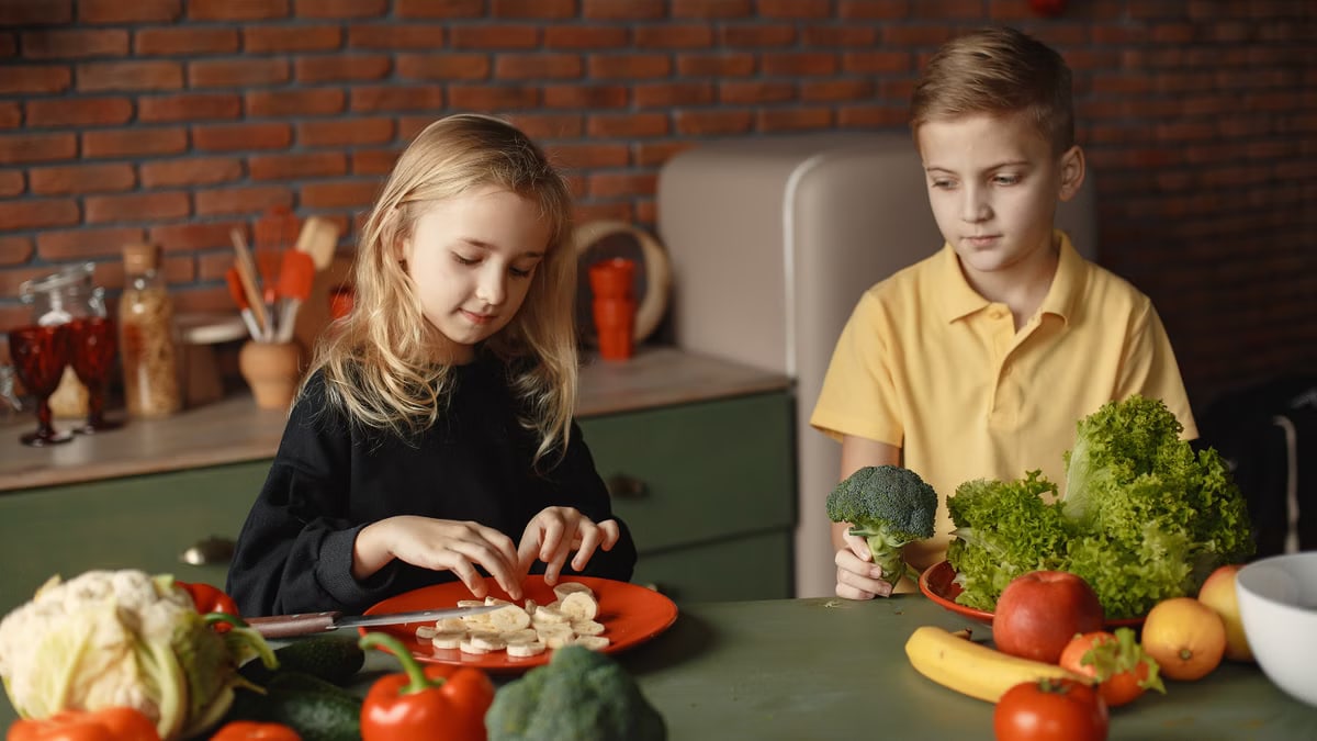 Positive little girl and boy sitting at desk with various fresh vegetables and fruits while preparing meal in loft kitchen with brick wall
